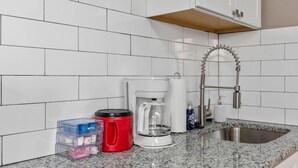 A kitchen with white tiled walls, a granite countertop, a stainless steel sink, a red kettle, a white coffee maker, and a plastic storage box. - The Lewis & Clark Hangout (Saint Charles)
