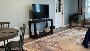 Una sala de estar con televisor sobre un mueble bajo, una mesa de comedor con sillas, un tapete estampado y un ventilador de techo.
