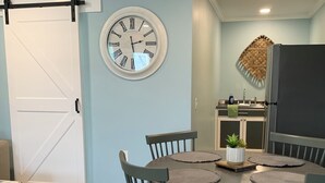 Un comedor con una mesa redonda, cuatro sillas, un reloj, una puerta y una zona de cocina con un refrigerador.