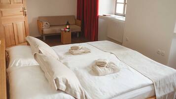 A neatly made bed with white linens and towels, a wooden wardrobe, a sofa, and a window with red curtains.