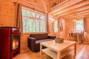 A cozy cabin interior with a wood-burning stove, a sofa, a dining table, and large windows offering a forest view.