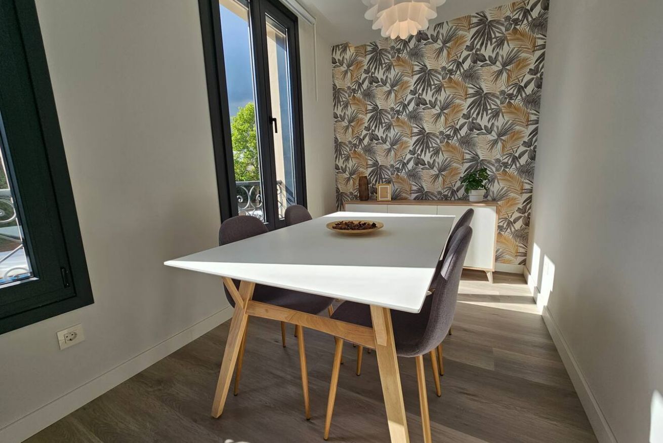 A dining area with a white table, wooden chairs, a small plant, and a patterned wall.