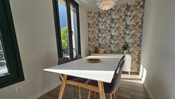 Ein Essbereich mit einem weißen Tisch, Holzstühlen, einer kleinen Pflanze und einer Wand mit Muster.