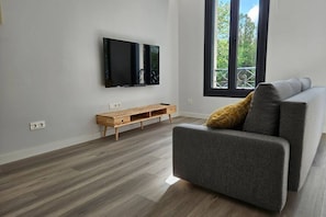 Ein modernes Wohnzimmer mit einem grauen Sofa, einer Holz-Unterhaltungsecke und einem großen Fenster mit Balkonblick.