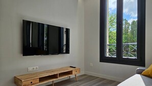 Ein modernes Zimmer mit einem großen, an der Wand montierten Fernseher, einer Holzbank und einem Fenster mit Balkonblick.