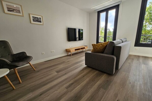 A modern living room with a grey sofa, a black chair, a wooden coffee table, and a flat-screen TV mounted on the wall.