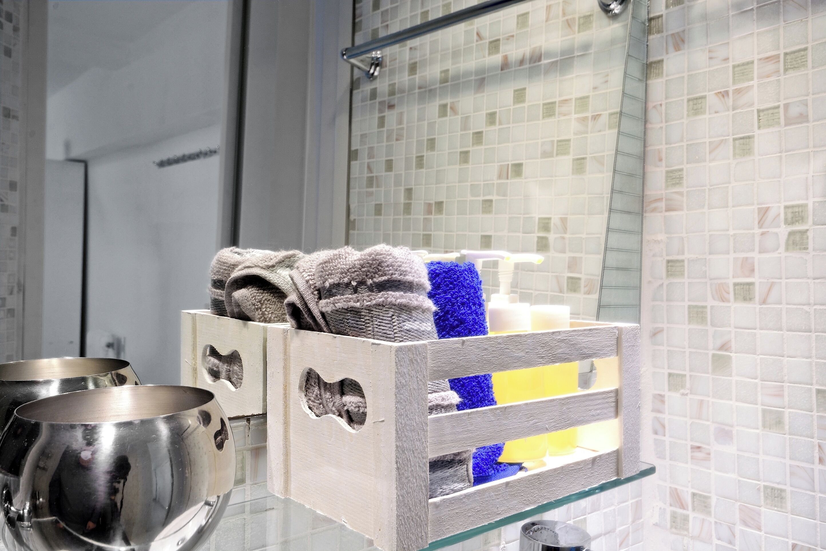 A bathroom with a glass shelf holding towels and toiletries, a mirror, and a tiled wall.