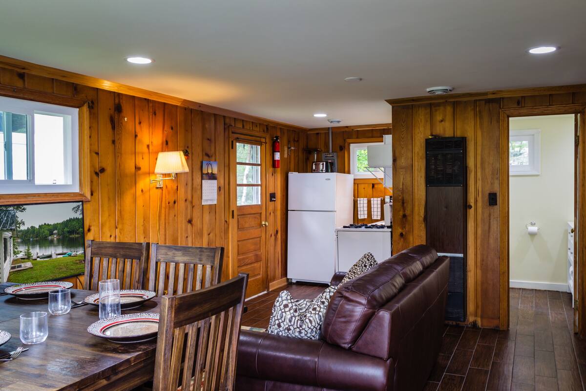 A cabin interior with a dining area, a living room with a leather couch, wooden walls, a refrigerator, and a window with a view of a lake.