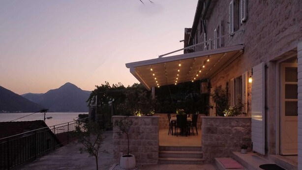 A covered outdoor patio with a table and chairs, stone planters, and a view of the sea and mountains at dusk.