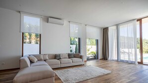 Un salón espacioso con un sofá seccional color beige, suelo de madera y grandes ventanales que ofrecen vistas a zonas verdes y a una piscina.
