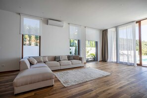 A spacious living room with a beige sectional sofa, a wooden floor, and large windows offering views of greenery and a pool.