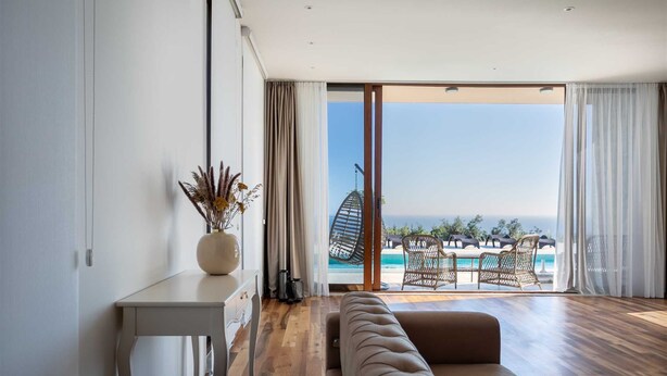 Una sala de estar moderna con piso de madera, un sofá color beige, una mesa consola blanca con un jarrón de flores secas y un amplio ventanal corredizo que da a un balcón con piscina y sillas de descanso.