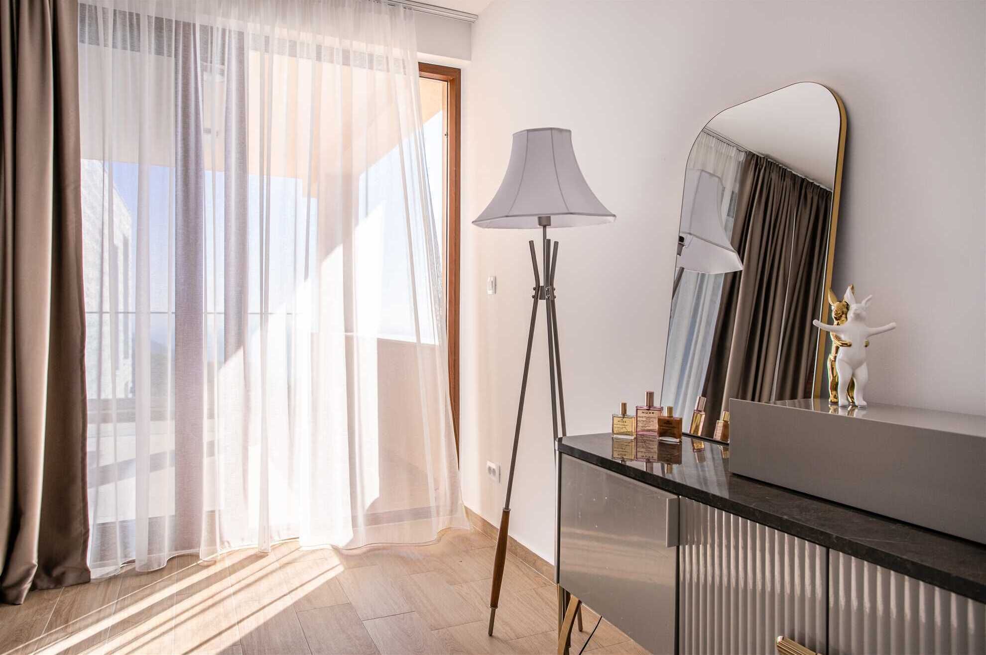 Une chambre à coucher moderne avec une grande fenêtre, des rideaux légers, une lampe sur pied, une coiffeuse avec un miroir et une petite statue.