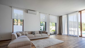 A spacious living room with a beige sectional sofa, a wooden floor, and large windows offering views of greenery and a pool. - Villa Beta with Pool (Budva)
