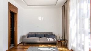 A modern living room with a grey sofa, wooden flooring, and a large window with sheer curtains. - Villa Gama with Pool (Unknown)