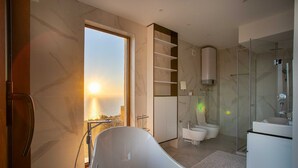 A modern bathroom with a freestanding bathtub, a glass shower enclosure, and a view of the sea through a large window.