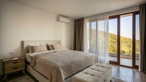 A bedroom with a large bed, a nightstand, an air conditioning unit, and a view of a hillside through a sliding glass door.