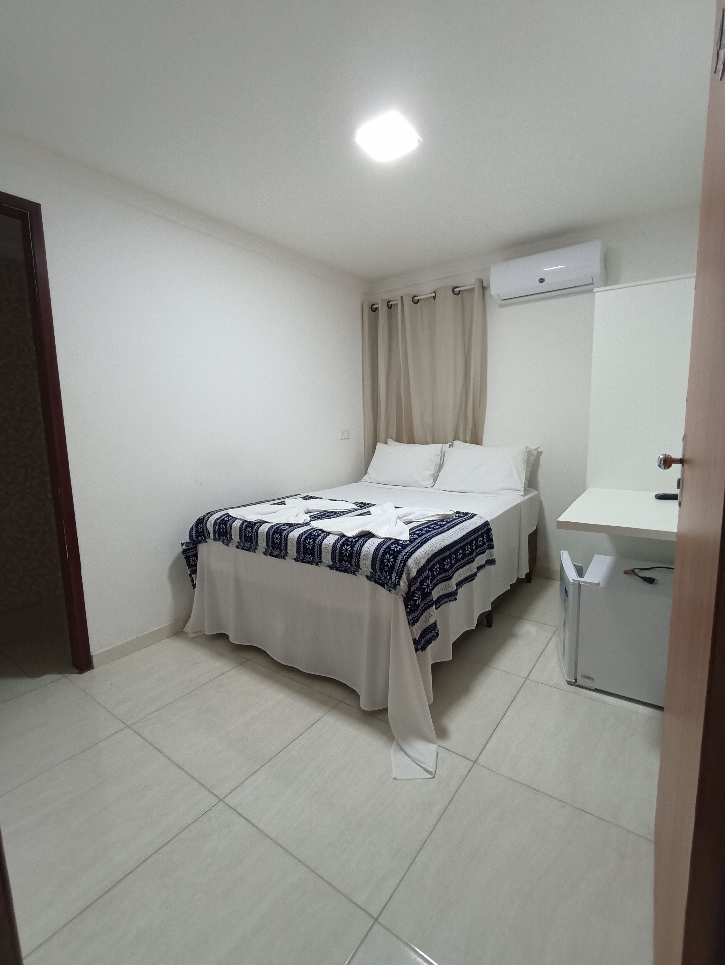 A small, clean room with a bed, a bedside table, a mirror, and a white air conditioning unit on the wall.