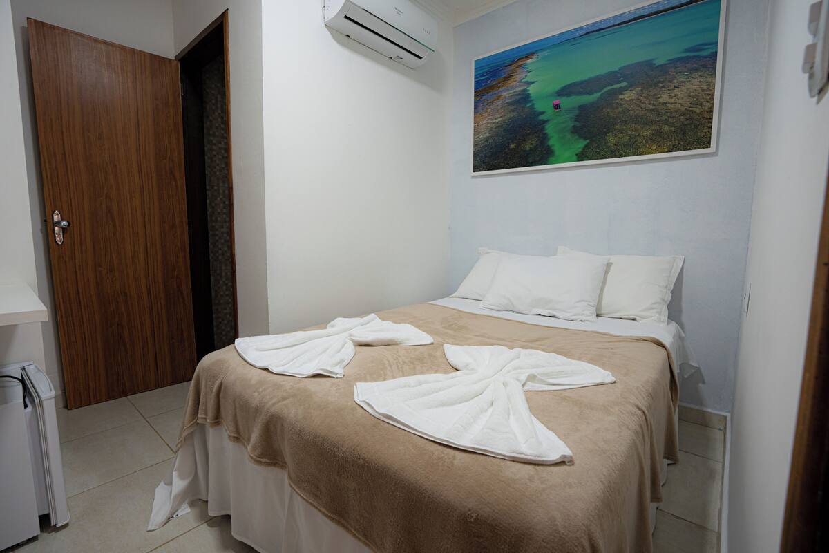 A hotel room with a bed, a painting on the wall, and an air conditioning unit.