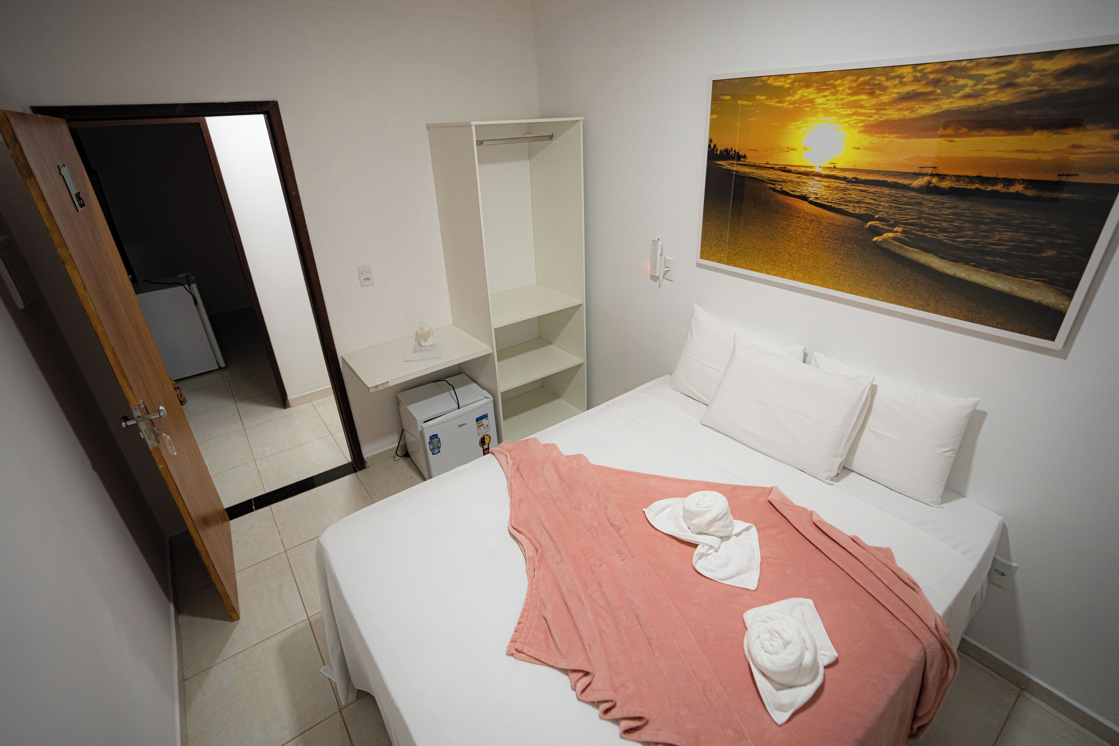 A hotel room with a bed, a white wardrobe, a small desk, and a painting of a sunset over the ocean.