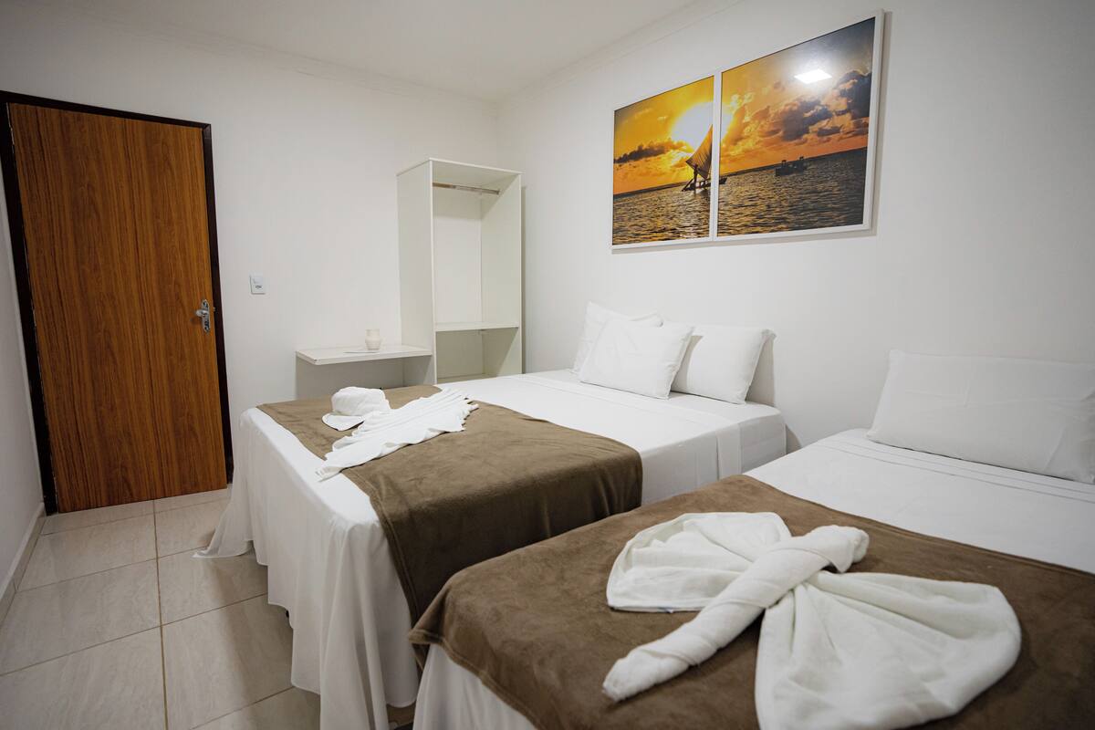 A hotel room with two beds, a wooden door, a white bedside table, a mirror, and a sunset painting on the wall.
