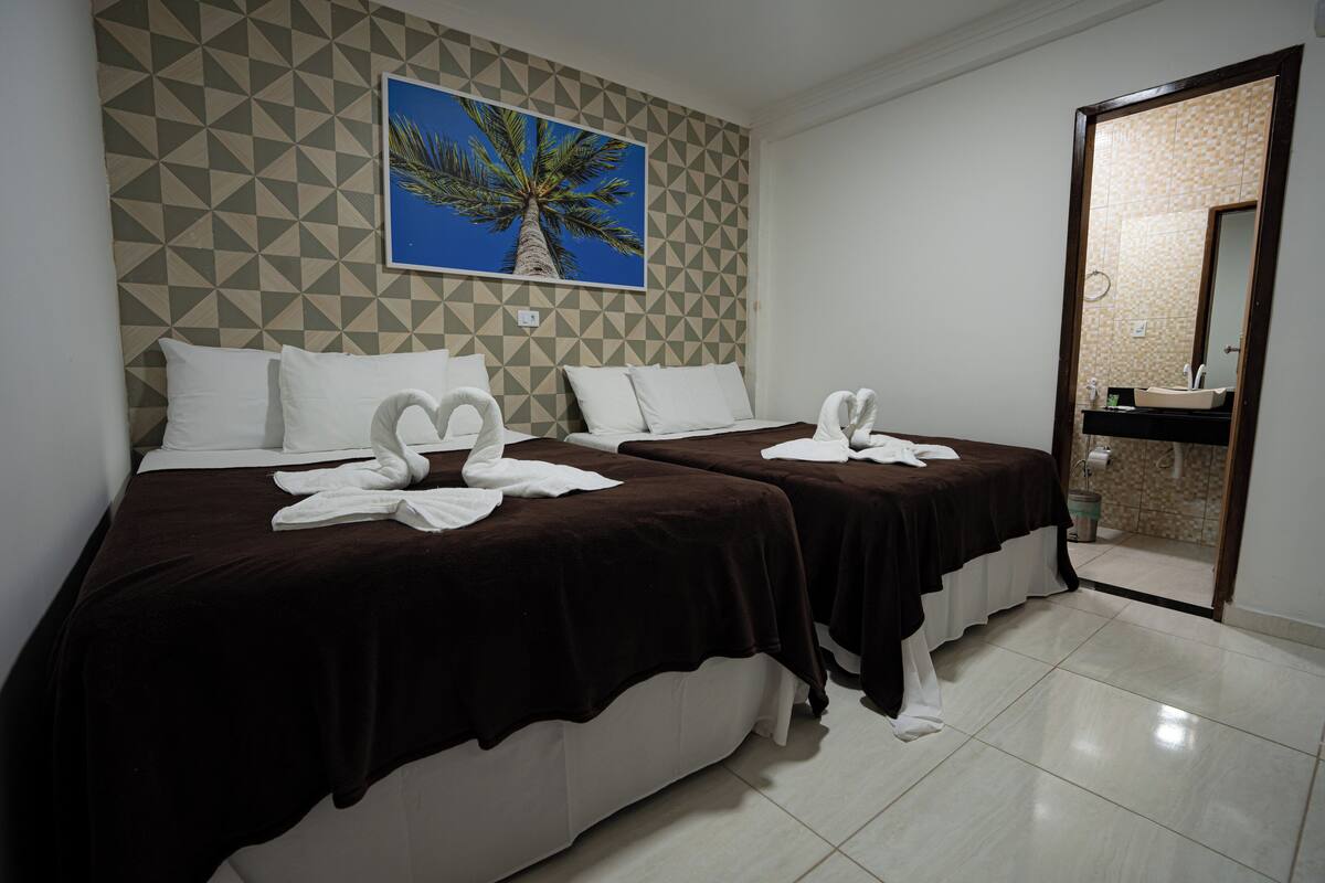 A hotel room with two beds, each with a brown bedspread and white pillows, and a framed picture of a palm tree on the wall.