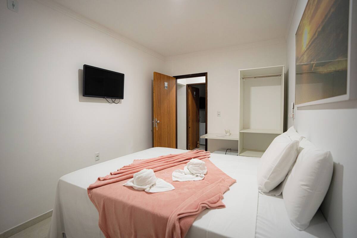 A hotel room with a bed, a television mounted on the wall, a wooden door, a mirror, and a shelf.