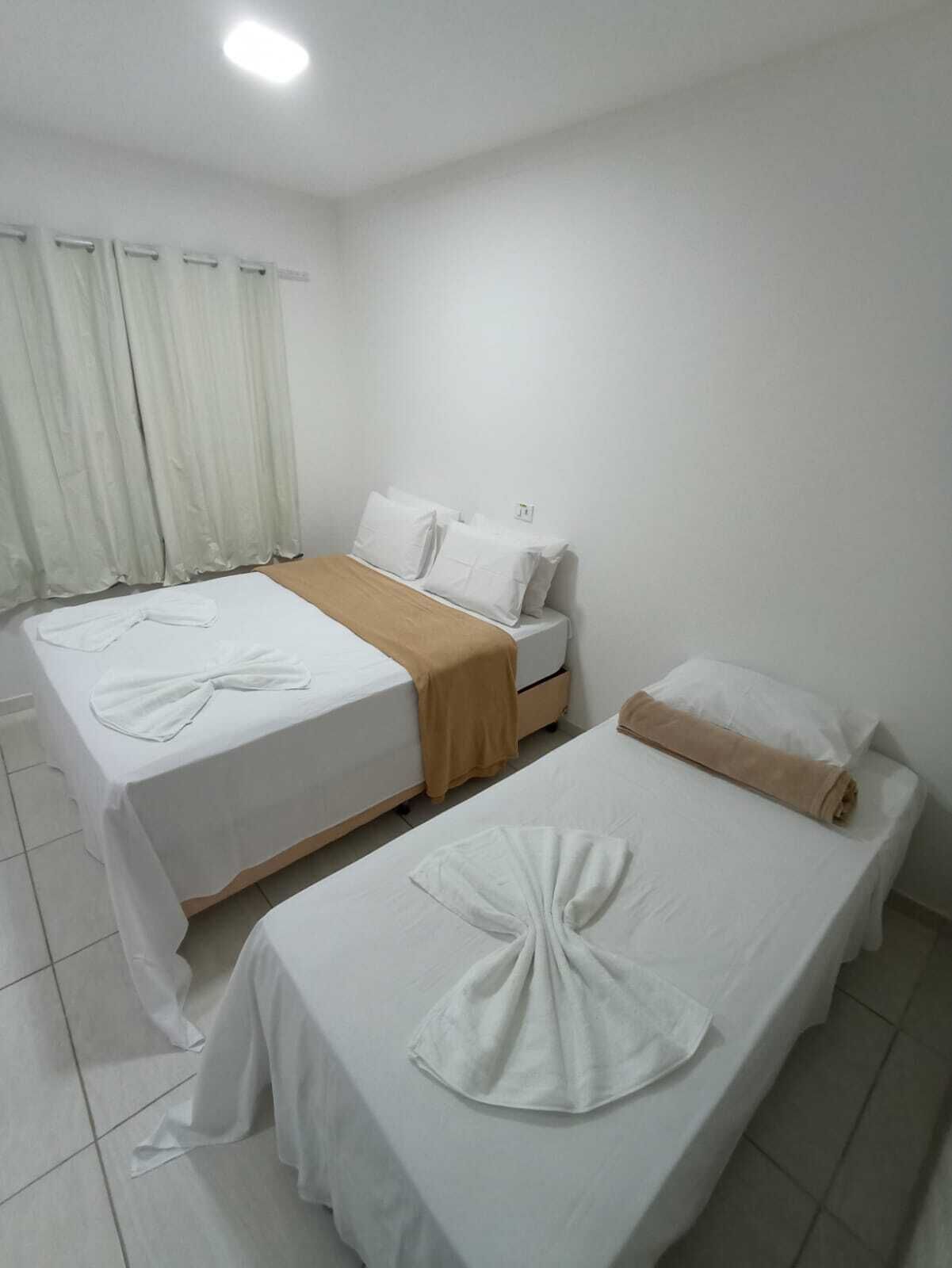 A hotel room with two beds, each with white linens and a towel. There is a beige throw blanket on one bed. The room has white curtains and a tiled floor.