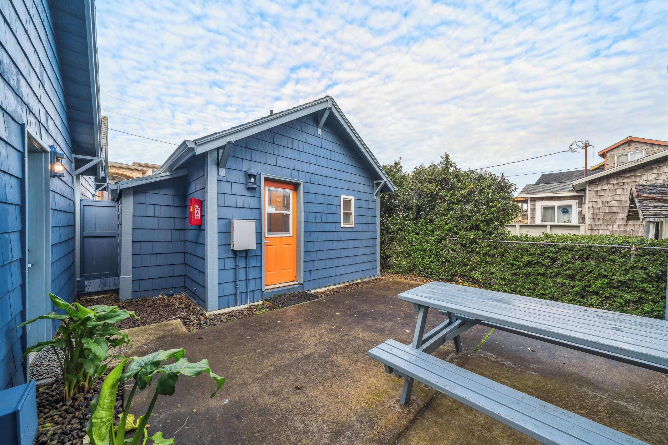 Een klein, blauw gebouw met een oranje deur, een picknicktafel en een haag op de achtergrond.