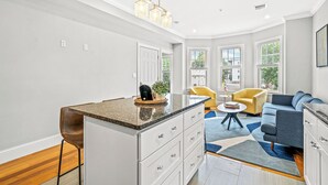 Et moderne køkken med en granitbordplade, hvide skabe og en indbygget ø. I det tilstødende opholdsområde er der en gul lænestol og en blå sofa.