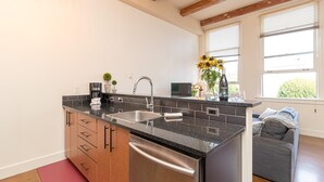 Eine moderne Küche mit einem Edelstahl-Geschirrspüler, einer integrierten Spüle und einer dunklen Granitarbeitsplatte. Auf der Arbeitsplatte stehen eine Kaffeemaschine, eine Vase mit Sonnenblumen und eine Weinflasche.