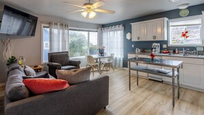 A modern living room with a sofa, a dining table, and a kitchen area with white cabinets and a window with flowers. - Blue Octopus #6 Beach Access. Ground Level. (Rockaway Beach)