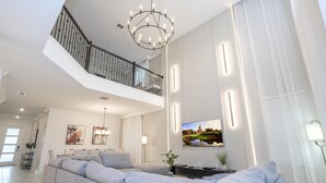 Een ruime woonkamer met een grote grijze modulaire bank, een kroonluchter, een flatscreen televisie aan de muur en een trap naar een hoger gelegen verdieping.