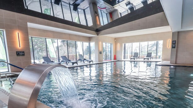 An indoor swimming pool with a waterfall feature, lounge chairs, and large windows. - Woodlands Retreat (Ruthin)