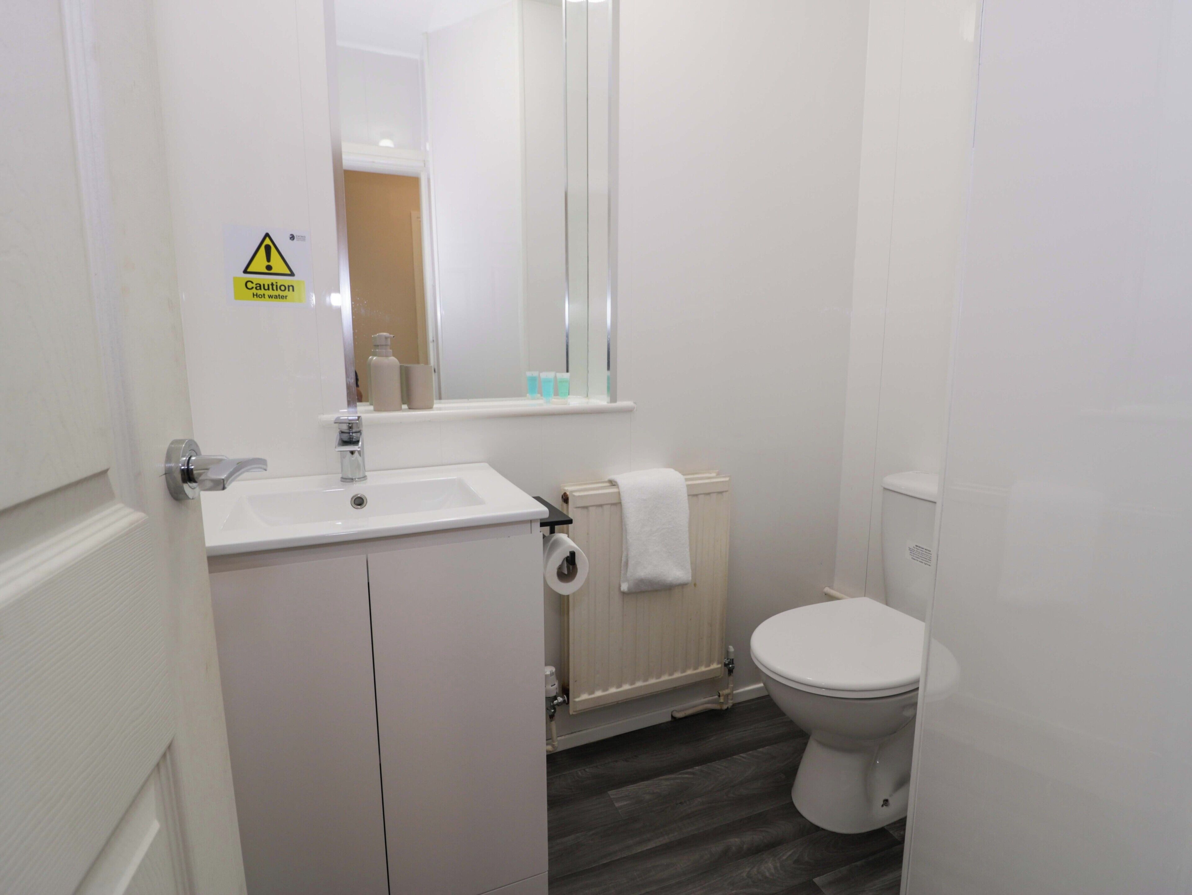 A bathroom with a white sink, a toilet, a mirror, and a caution sign for hot water.