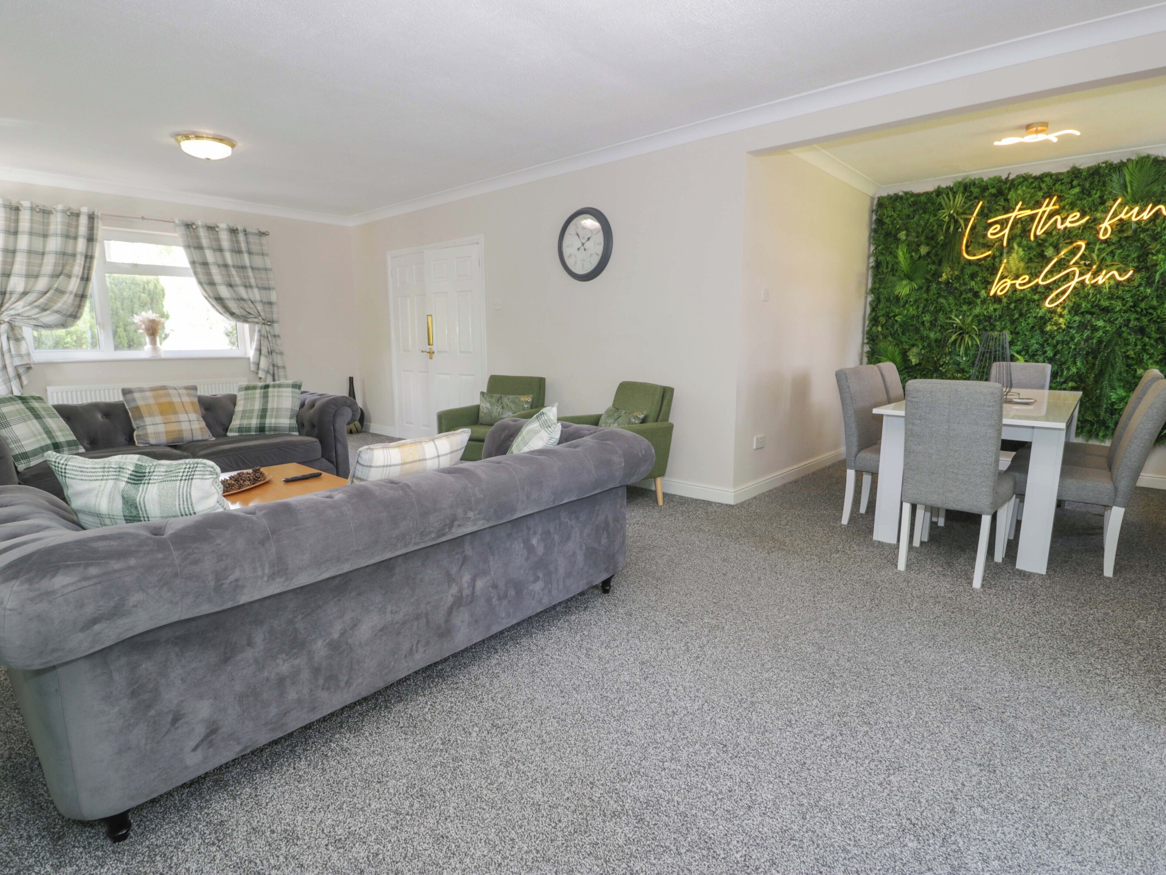 Een woonkamer met een grijze bank, een eettafel met stoelen, een groene fauteuil, een klok aan de muur en een neonbord.