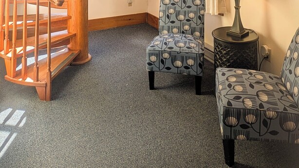 Una habitación con una escalera espiral de madera, dos sillones estampados, una mesita con una lámpara y una ventana con cortinas.