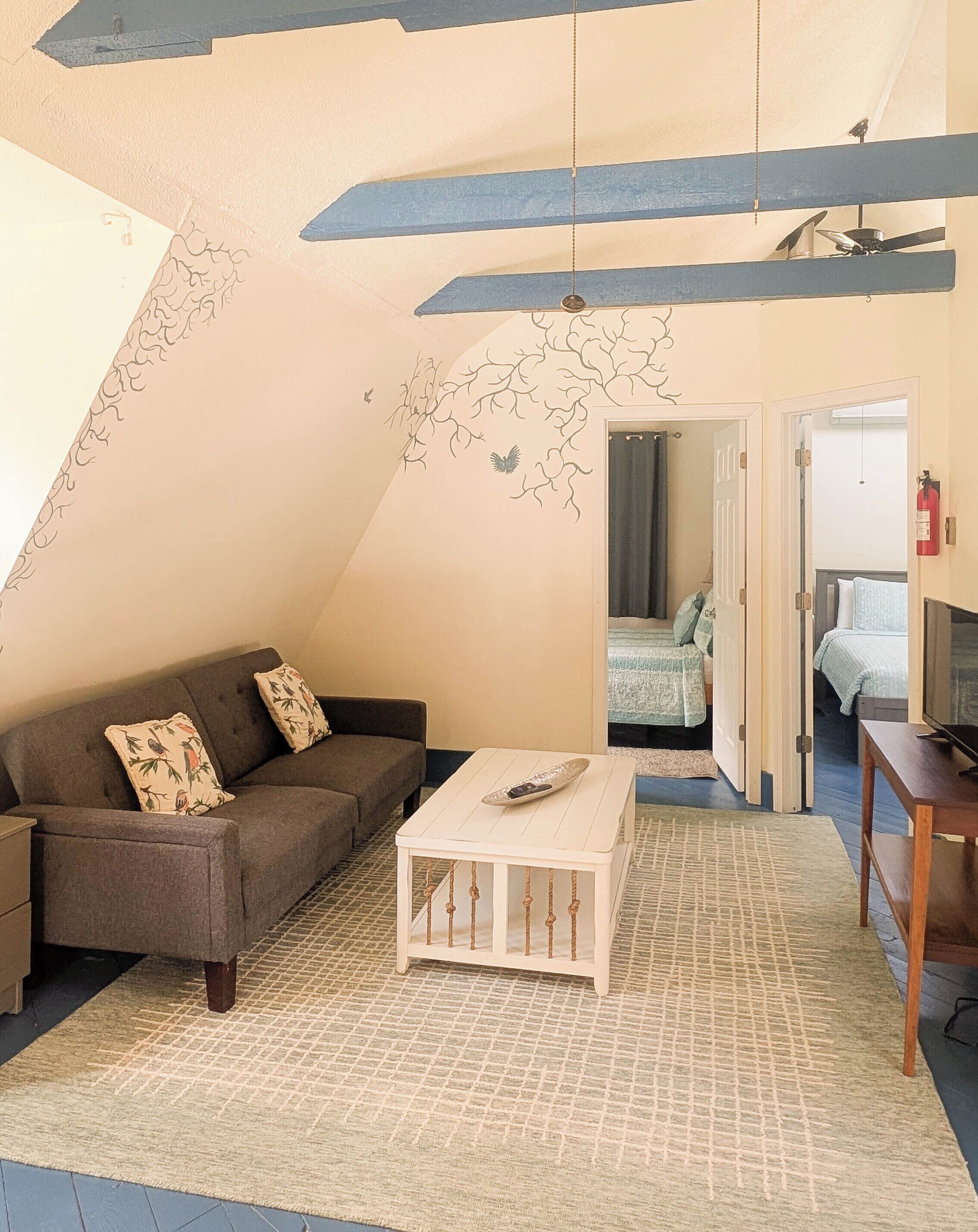 Une chambre à coucher confortable, aménagée en mezzanine avec un plafond en pente, un canapé agrémenté de coussins à motifs, une petite table blanche et un meuble pour la télévision.