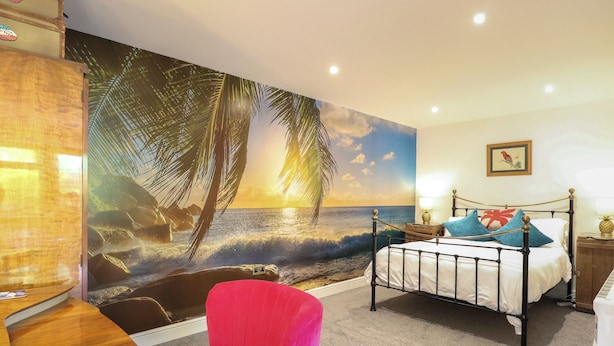 Une chambre à coucher avec un lit, un fauteuil rouge, un bureau en bois et une grande fresque murale représentant une scène de plage.