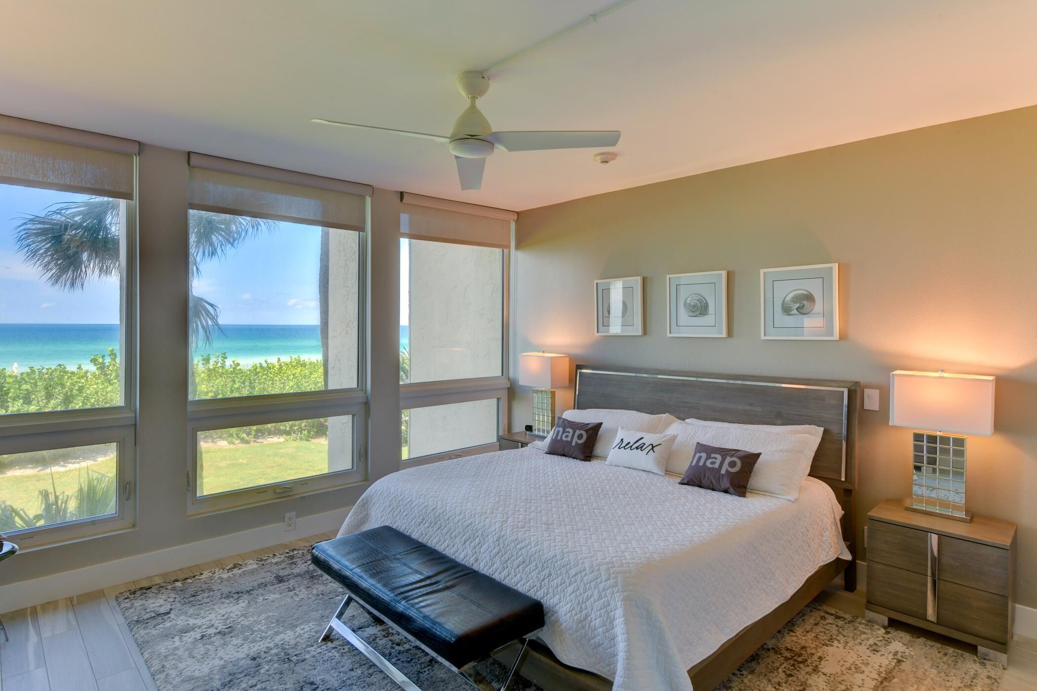 A bedroom with a large bed, a bench, a ceiling fan, and a view of the ocean.