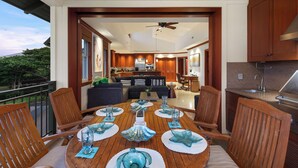 A dining area with a wooden table set for six, surrounded by wooden chairs. A kitchen is visible in the background with wooden cabinets and a stainless steel sink. - Hilton Pool Pass Kolea - Luxe Penthouse (Waikoloa)