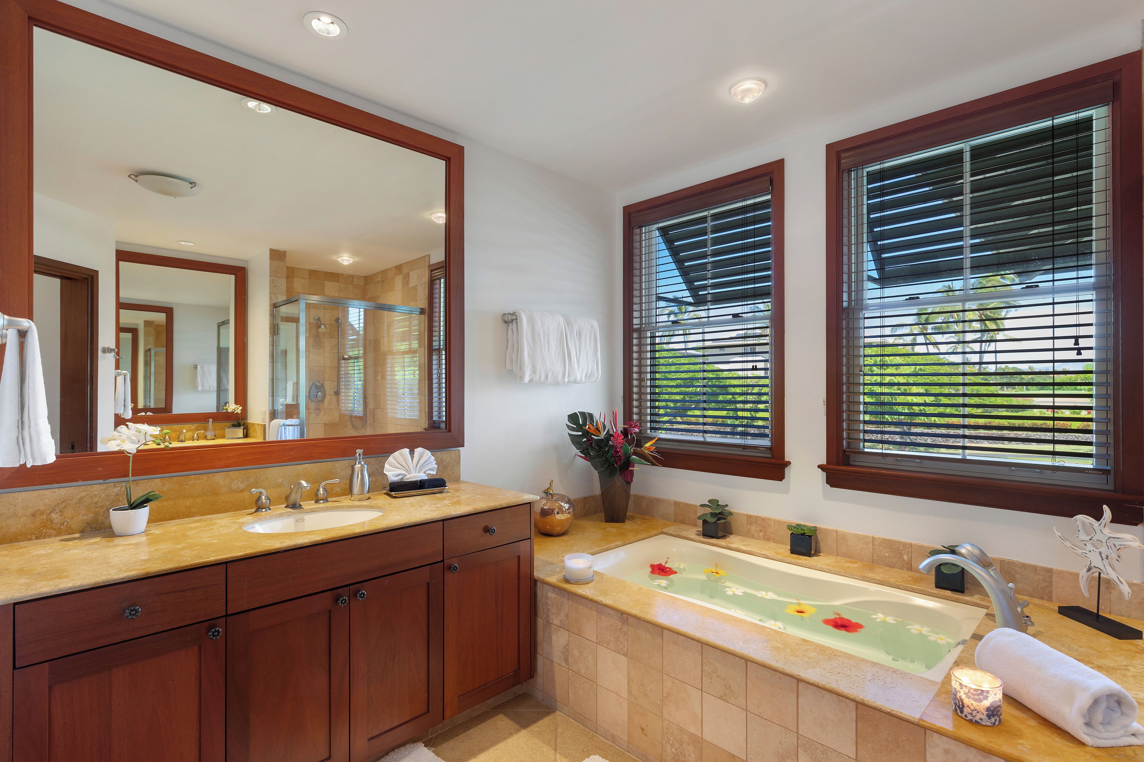 Un baño moderno con un espejo grande, lavamanos dobles, una bañera con un arreglo floral y vistas a zonas verdes a través de las ventanas.