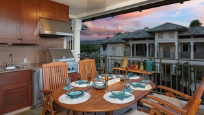 A dining area with a wooden table set for four, a kitchen with modern appliances, and a balcony with a view of houses and a sunset. - Hilton Pool Pass Kolea - Luxe Penthouse (Waikoloa)