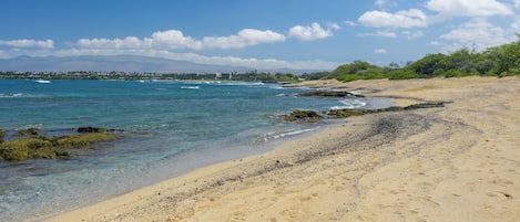 A sandy beach with clear water, a rocky shoreline, and distant mountains.
