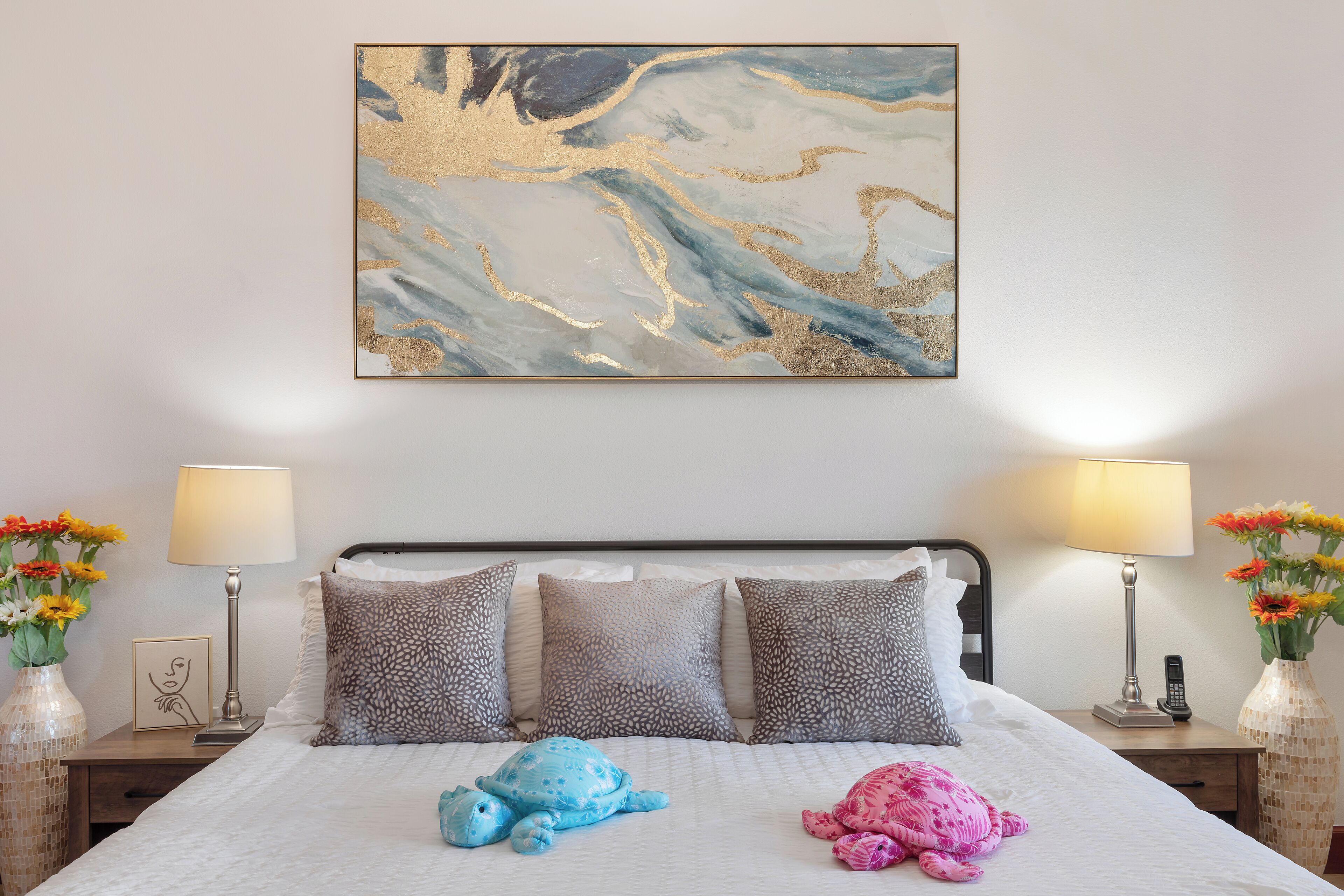 Una cama bien tendida con almohadones estampados, un peluche azul y rosa, y un cuadro abstracto colgado en la pared.