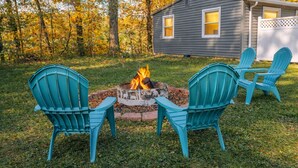 Un jardin avec un foyer entouré de fauteuils Adirondack, une terrasse en bois et une maison en arrière-plan.
