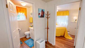A compact room with a toilet, a bed with a yellow bedspread, a blue mat, and a coat rack.