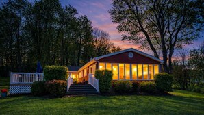 A two-story house with a front porch and a deck, surrounded by trees and a well-maintained lawn. - Lake Front Oasis: 3 Bed 2 Ba Cottage on Lake Logan (Logan)