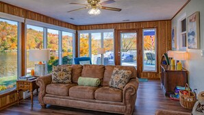 A cozy living room with a brown leather sofa, a wooden floor, and large windows offering a view of a lake and autumn foliage. - Lake Front Oasis: 3 Bed 2 Ba Cottage on Lake Logan (Logan)