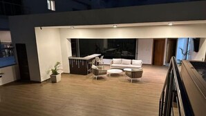 A modern living area with a sofa, coffee table, and chairs. There is a bar area with a counter and seating. The space has a wooden floor and large windows.